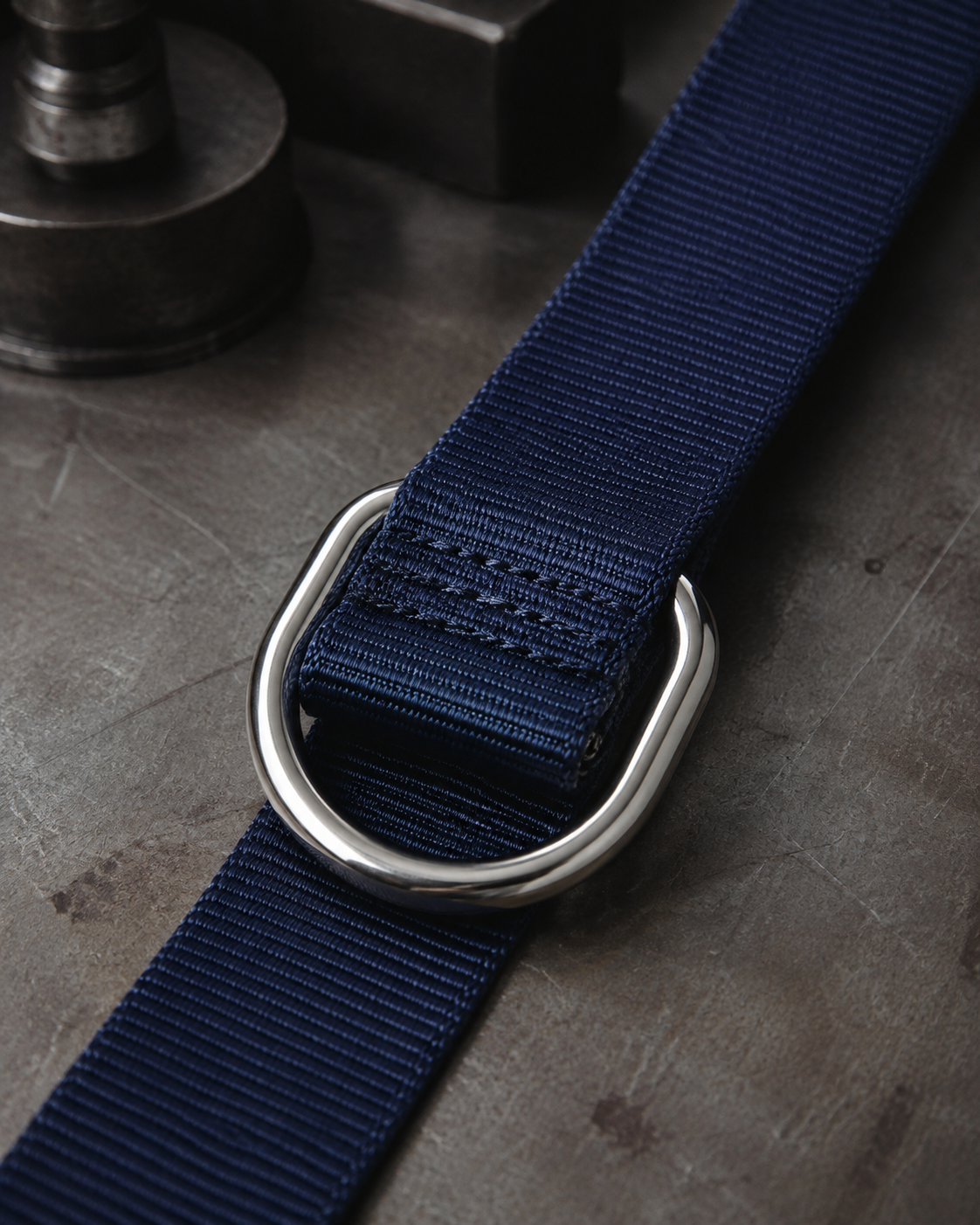 Navy nylon webbing threaded through a polished steel D-ring on a factory workbench.